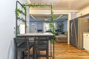 Una cocina con encimera de mármol, dos sillas, un refrigerador y una zona de comedor con ventilador de techo.