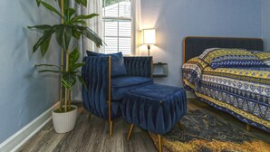 Ein Zimmer mit einem blauen Sofa, einer gemusterten Bettdecke, einer Topfpflanze und einer Lampe.
