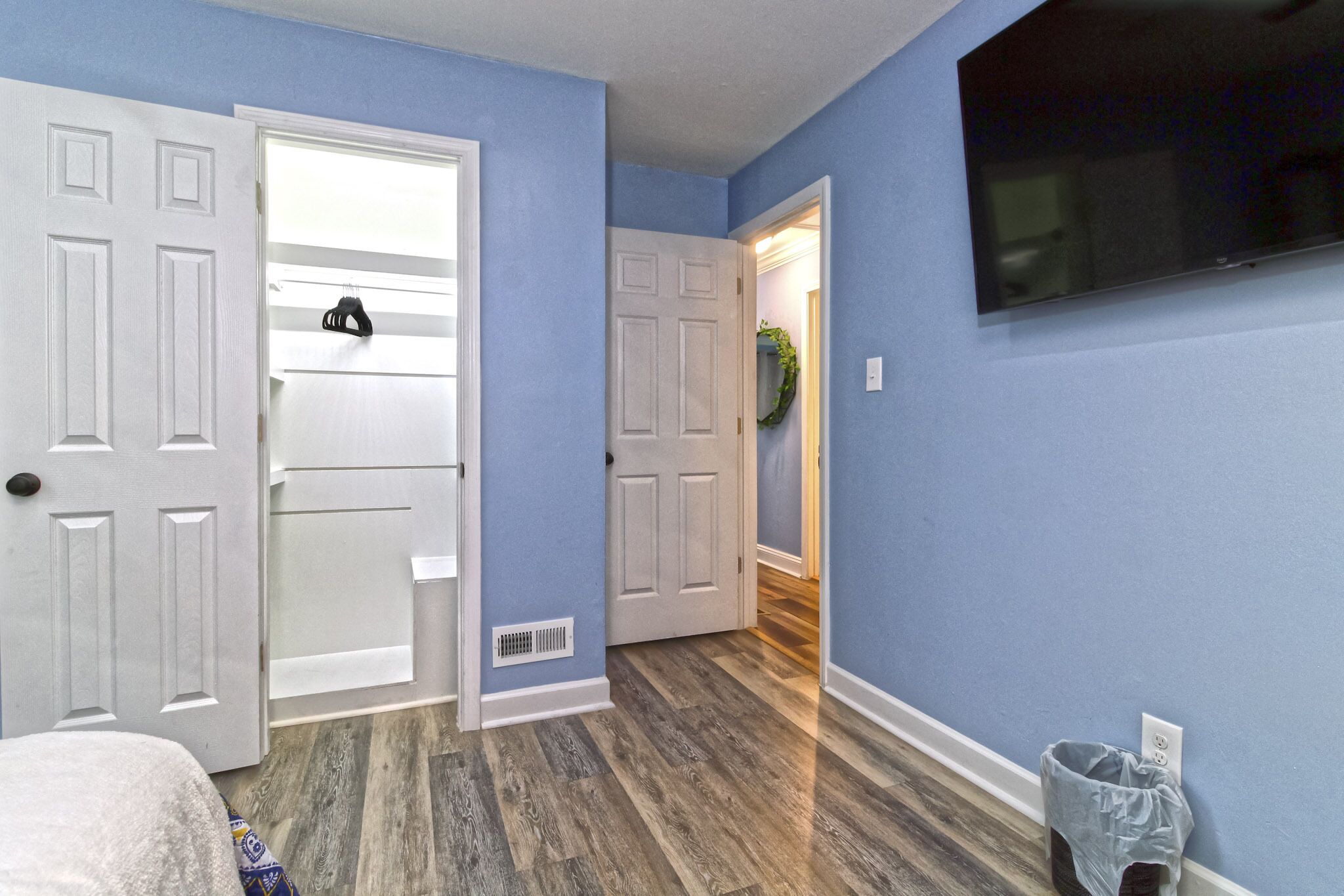 A room with blue walls, a wooden floor, a bed, a TV mounted on the wall, and a door leading to another room.