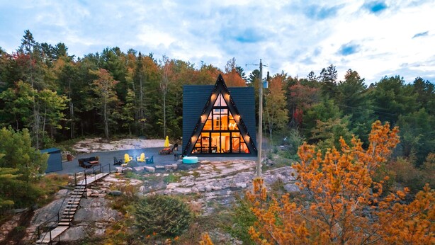 A cabin with a triangular roof and large windows, surrounded by autumn foliage and a rocky landscape. - Waterfront Timber A-Frame Cabin on French River (French River)