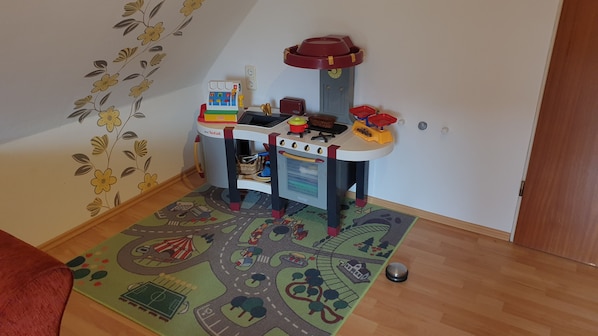 Un set de cocina infantil instalado en una habitación con diseño floral en la pared, un tapete de juego colorido y suelo de madera.
