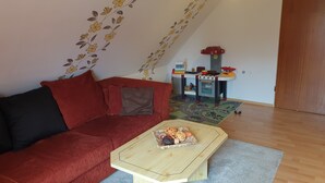 Una sala de estar acogedora con un sofá rojo, una mesa de centro de madera con aperitivos, una cocina de juguete para niños y una alfombra estampada.