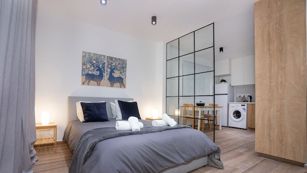 A modern bedroom with a large bed, a bedside table, a wall-mounted artwork, and a view into the adjacent kitchen area.