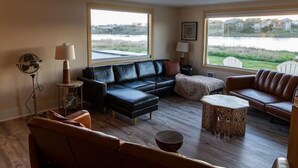 A spacious living room with a black leather sofa, a brown leather couch, and a round wooden coffee table. There are two windows offering a view of a body of water and a distant landscape. - Pacific City Riverfront Bungalow on Nestucca River! Dog friendly-Hot tub-Firepit (Pacific City)