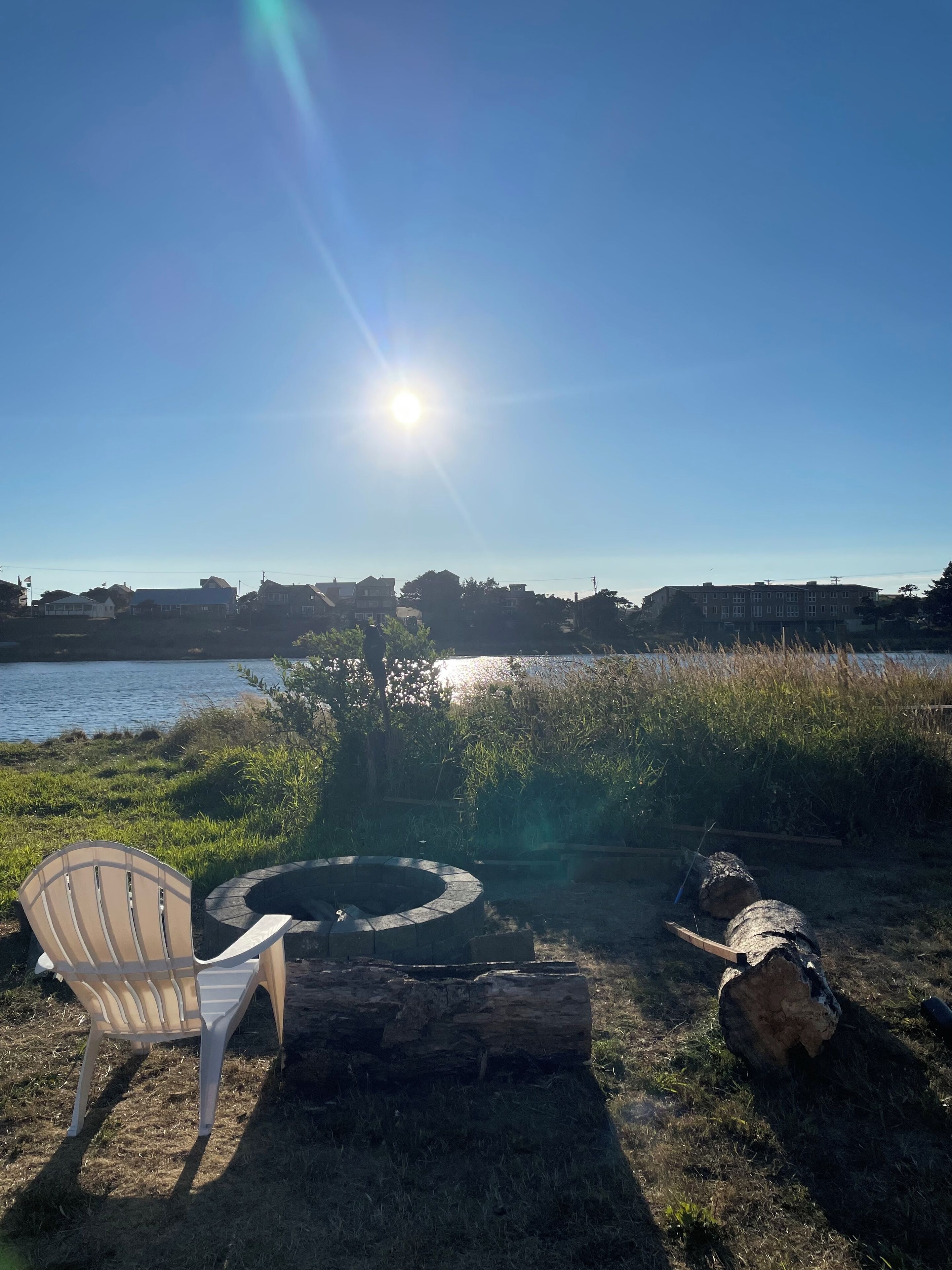 A clear sky with the sun shining, a body of water, and a fire pit with logs and a chair in the foreground.