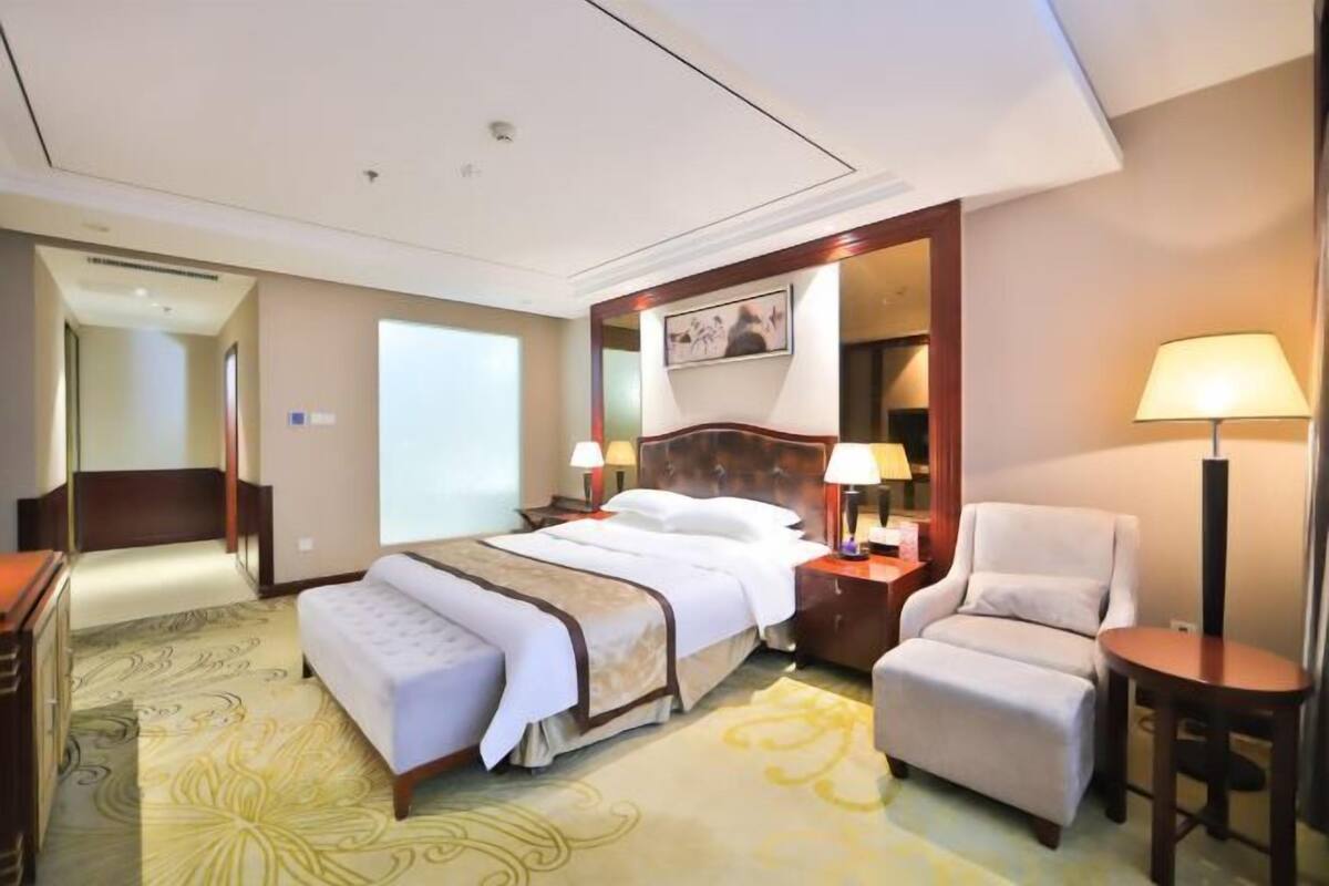 A hotel room with a large bed, a sitting area with a chair and a table, a desk, and a wardrobe.