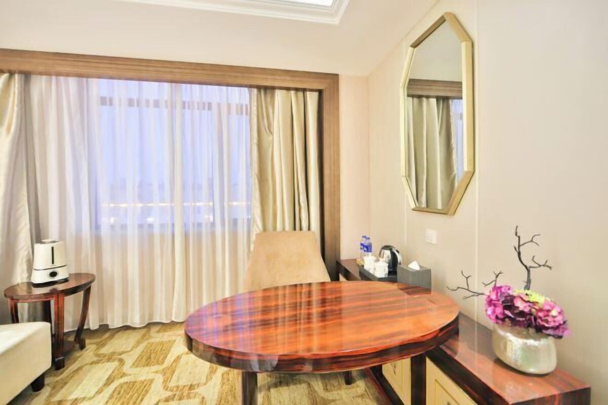 A hotel room with a round wooden table, a chair, a bed, a small table with a lamp, a mirror, and a vase with flowers.