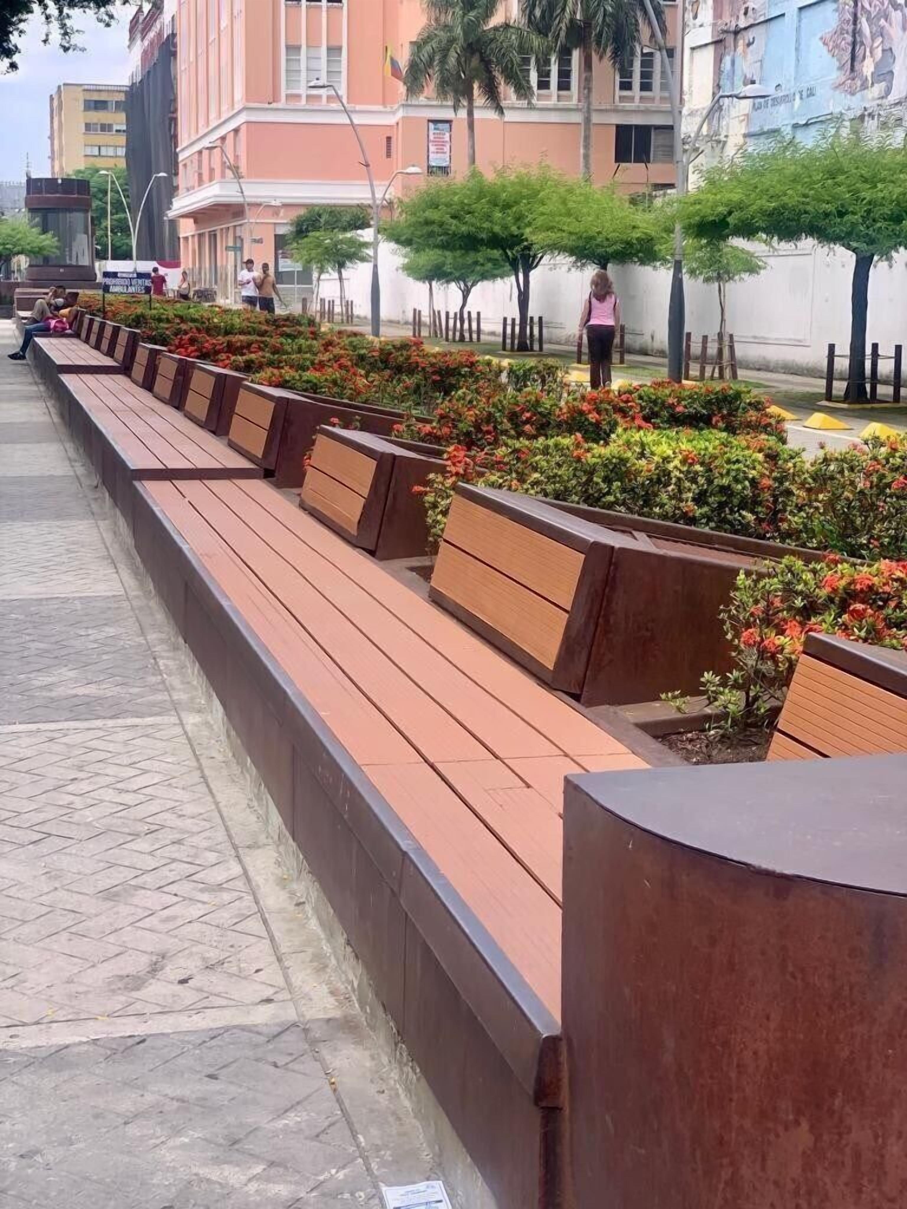 Una fila de bancos de madera a lo largo de un sendero pavimentado, con un jardín ajardinado que incluye arbustos y árboles pequeños.