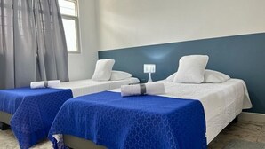 A hotel room with two beds, each with a blue bedspread and white pillows. There is a nightstand with a lamp between the beds. - Casa Nawi (Cali)