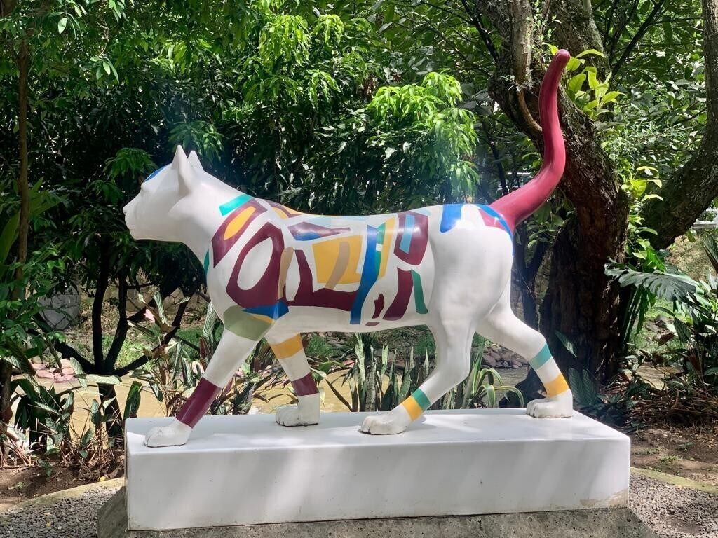 A colorful sculpture of a cat on a pedestal in a garden setting.