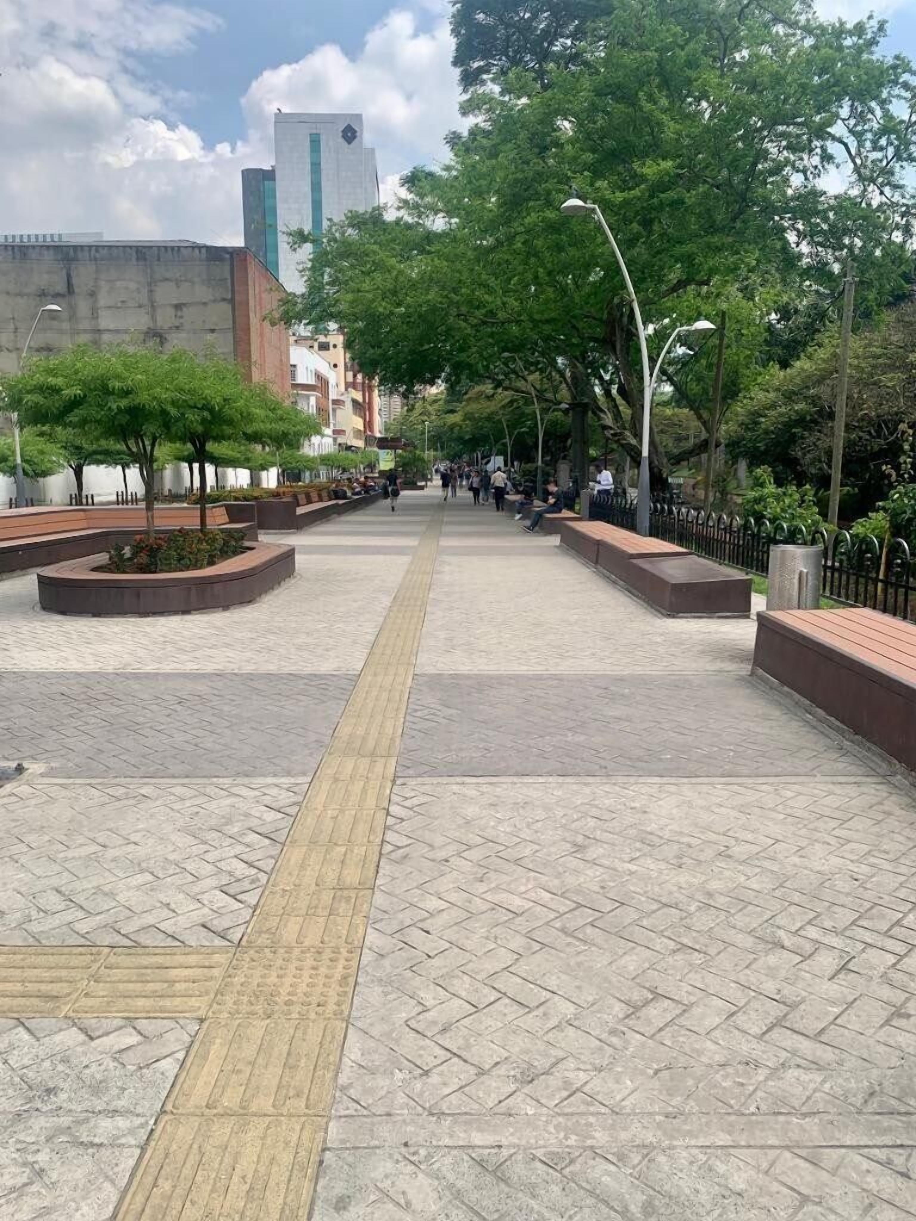 A spacious pedestrian area with a tactile paving path, surrounded by trees and benches.