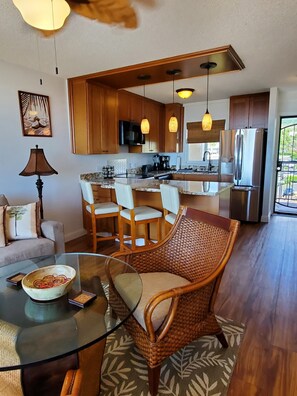 A modern kitchen with wooden cabinets, a stainless steel refrigerator, and a dining area with wicker chairs and a glass table.