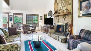 A living room with a stone fireplace, a television, and a dining area with a table and chairs. - Enjoy this cozy home with a wrap around porch, near the Smoky Mountains (Townsend)