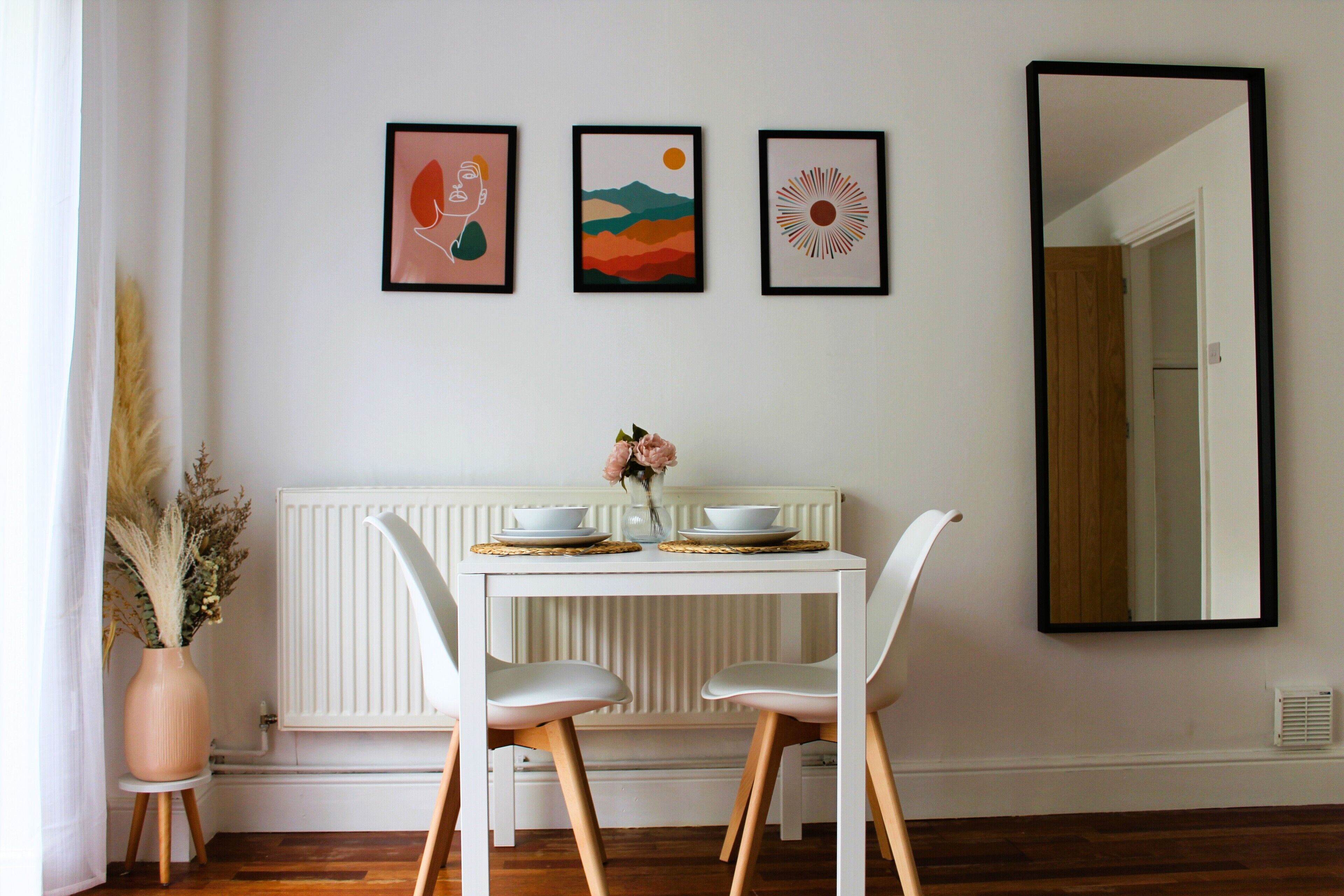 A dining area with a white table, two white chairs, a vase with flowers, and three framed artworks on the wall.
