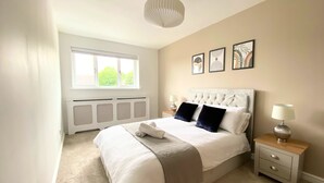A bedroom with a large bed, a window, a radiator, a bedside table with a lamp, and framed artwork on the wall.