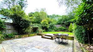 A patio area with a table and chairs, surrounded by stone tiles and greenery.