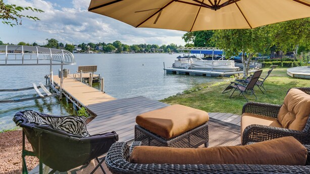 A waterfront patio with wicker furniture, a large umbrella, and a view of the lake and docked boats. - Okauchee Lake Vacation Rental w/ Boat Dock! (Oconomowoc)