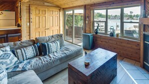 A cozy cabin interior with a wooden ceiling, a sofa with patterned cushions, a wooden coffee table, and a large window overlooking a lake. - Okauchee Lake Vacation Rental w/ Boat Dock! (Oconomowoc)