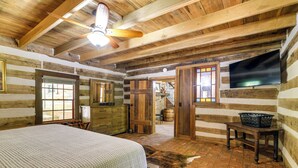 A rustic log cabin bedroom with a bed, wooden dresser, and a television mounted on the wall. - Glasgow Getaway w/ Patio, Fireplace & Grill! (Glasgow)