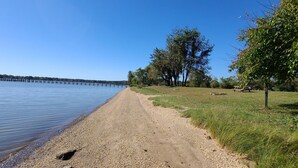 Ein Kiesstrand mit einer hölzernen Pier, die sich über ruhiges Wasser erstreckt, umgeben von Bäumen und Grünflächen.