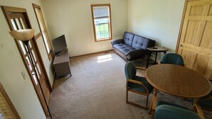 Ein Zimmer mit einer Couch, zwei Sesseln, einem kleinen Tisch, einem Fernseher auf einem Schrank, einem Fenster mit Jalousien und einer Holztür.