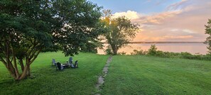 A grassy area with a stone path leading to a seating arrangement under trees, overlooking a body of water.