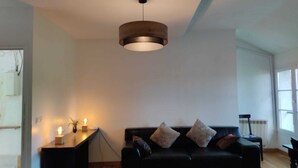 Una sala moderna con un sofá negro, una mesa de centro de madera, un lámpara colgante y un ventanal que proporciona luz natural.