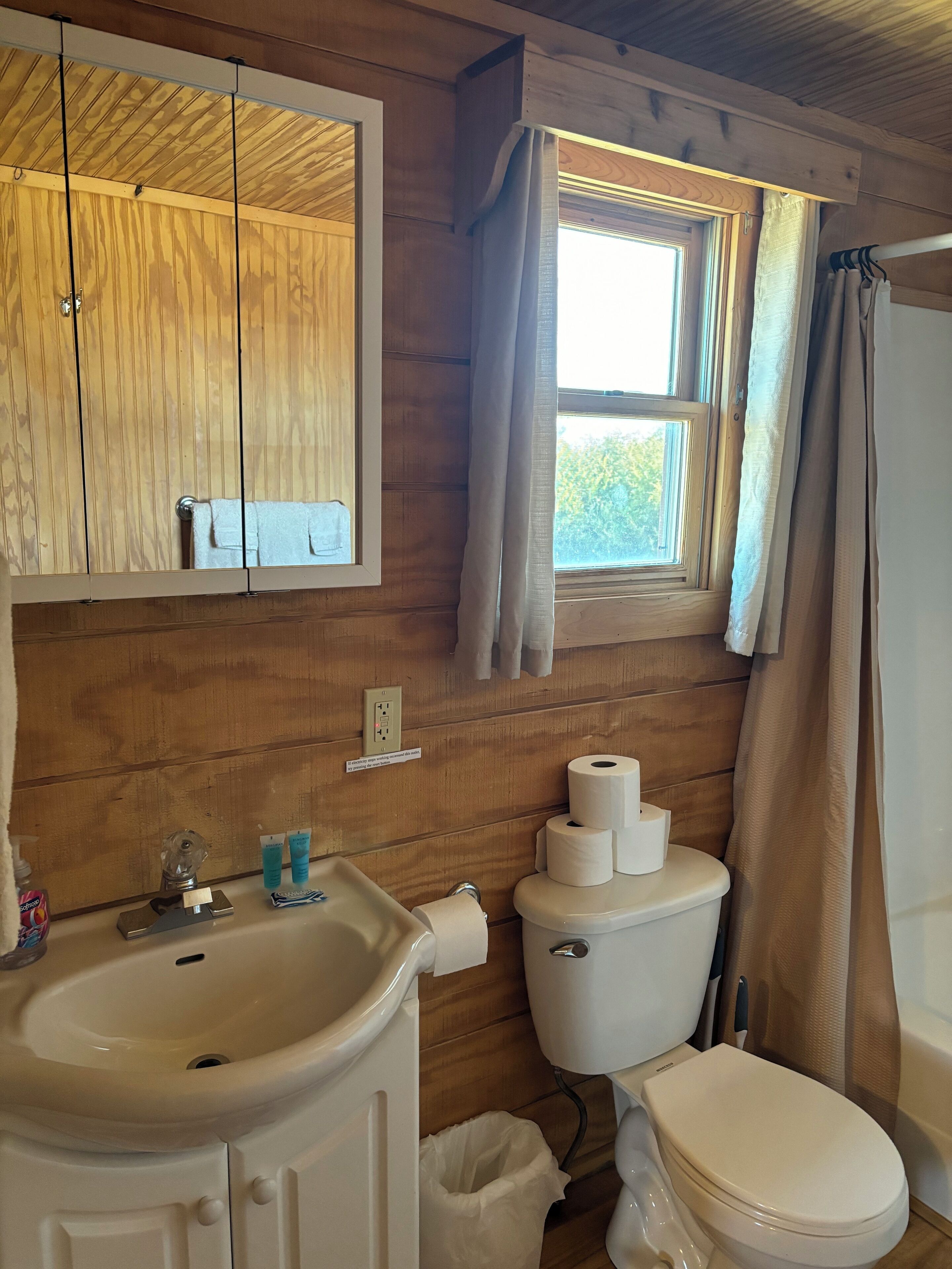 A wooden-paneled bathroom with a sink, mirror, toilet, and window with curtains.