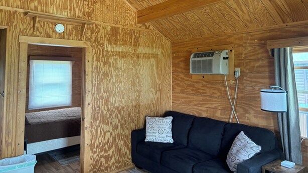 A wooden cabin interior with a black sofa, two patterned pillows, a ceiling fan, and a window with curtains. - Cabin close to Lake Rathbun #2
 (Melrose)