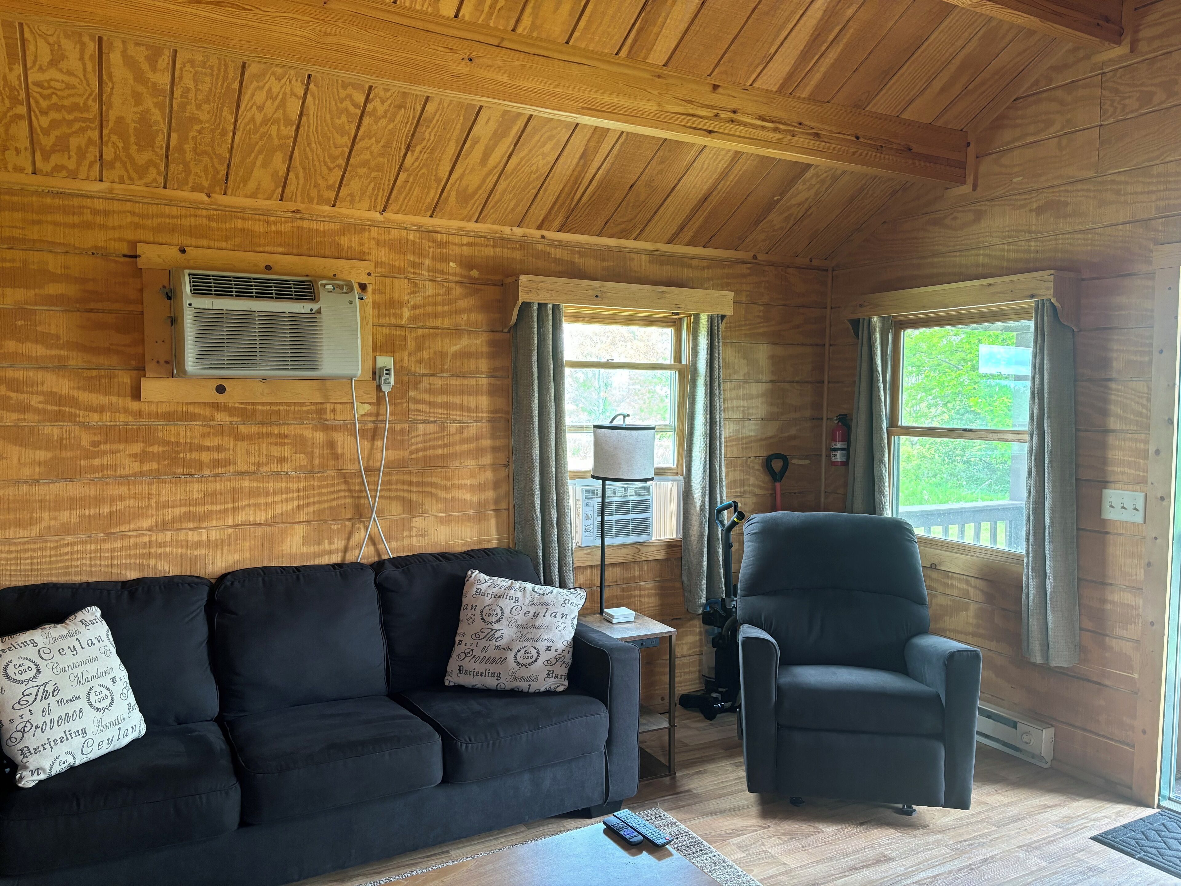 Ett mysigt rum med ett träpanelat tak, en svart soffa med mönstrade kuddar, en grå fåtölj och ett litet bord med en lampa.