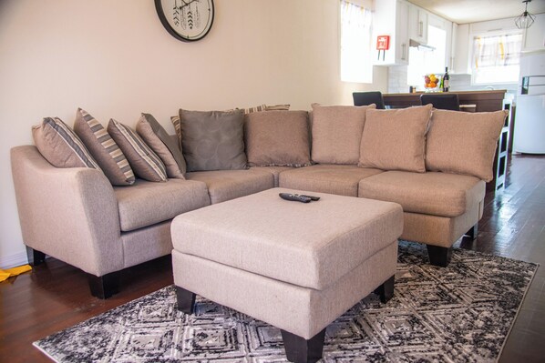 A living room with a beige sectional sofa, a matching ottoman, and a patterned rug.