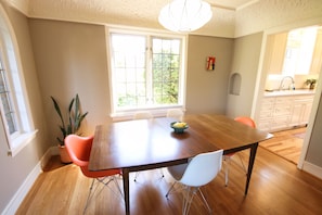 A dining area with a wooden table, orange chairs, and a bowl of fruit. There is a window with a view of trees and a kitchen in the background.