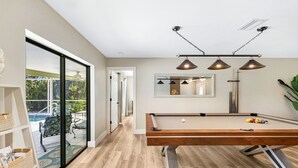 A pool table in a room with a wooden floor, a large window, and a ceiling-mounted lamp. - Billiards and Poolside Bliss Centrally located in SE Cape Coral, Pet Friendly! (Cape Coral)