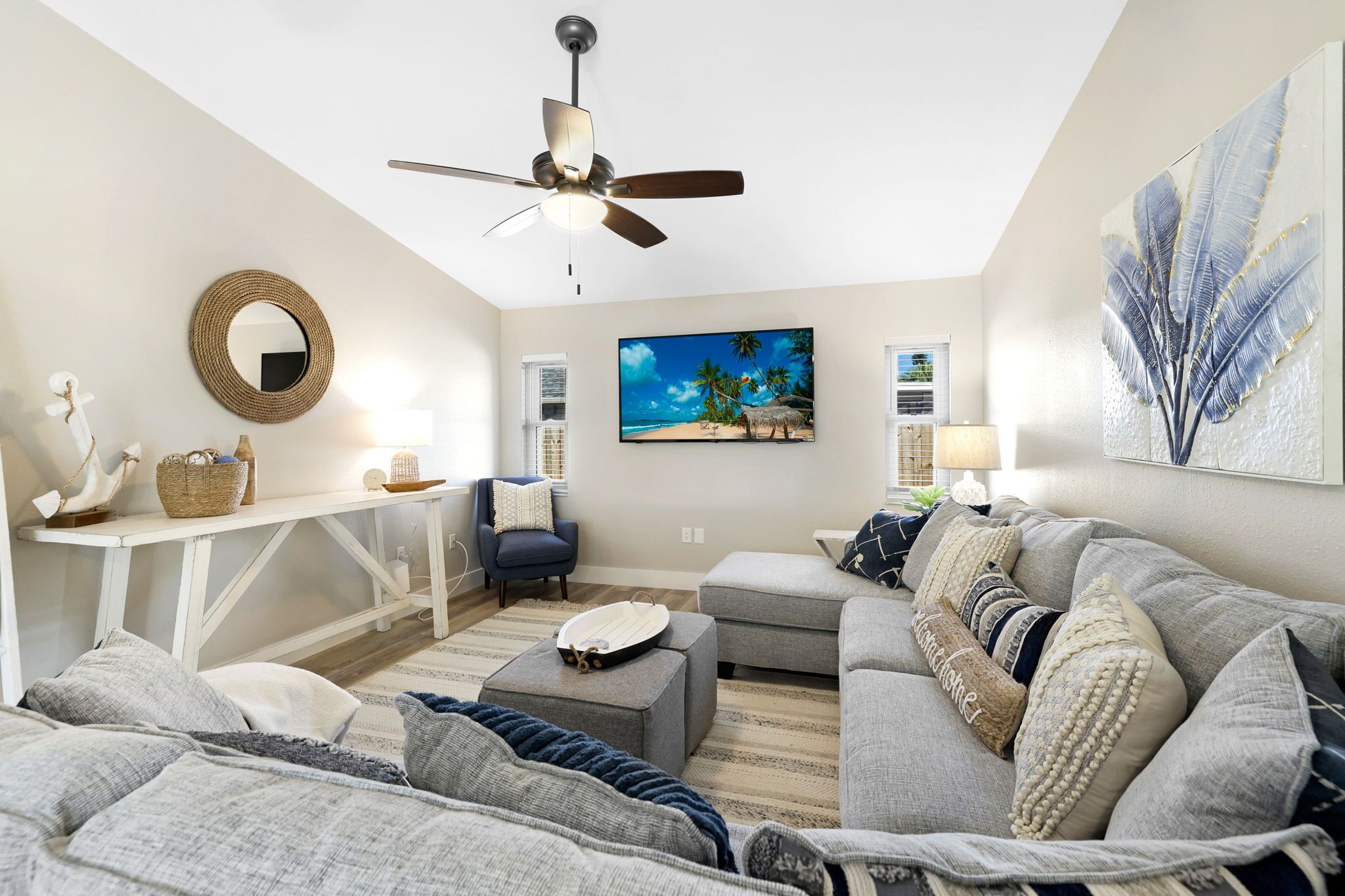 Een woonkamer met een plafondventilator, een groot schilderij van een strandlandschap, een ronde spiegel en een bank met meerdere kussens.