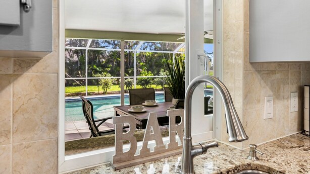 A kitchen with a marble countertop, a stainless steel faucet, and a window overlooking a pool and outdoor seating area. - Billiards and Poolside Bliss Centrally located in SE Cape Coral, Pet Friendly! (Cape Coral)