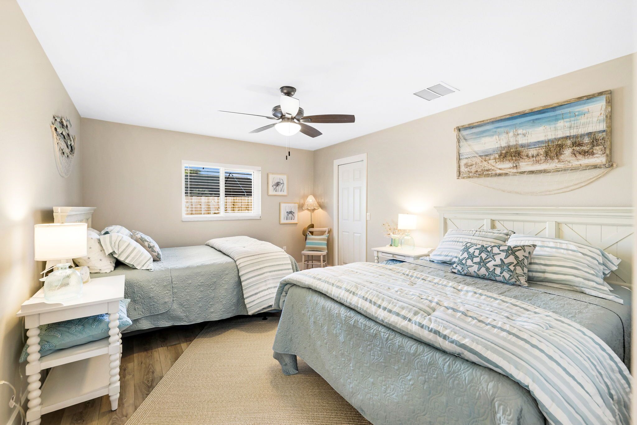 A bedroom with two beds, a ceiling fan, a window with blinds, a bedside table with a lamp, and a painting on the wall.