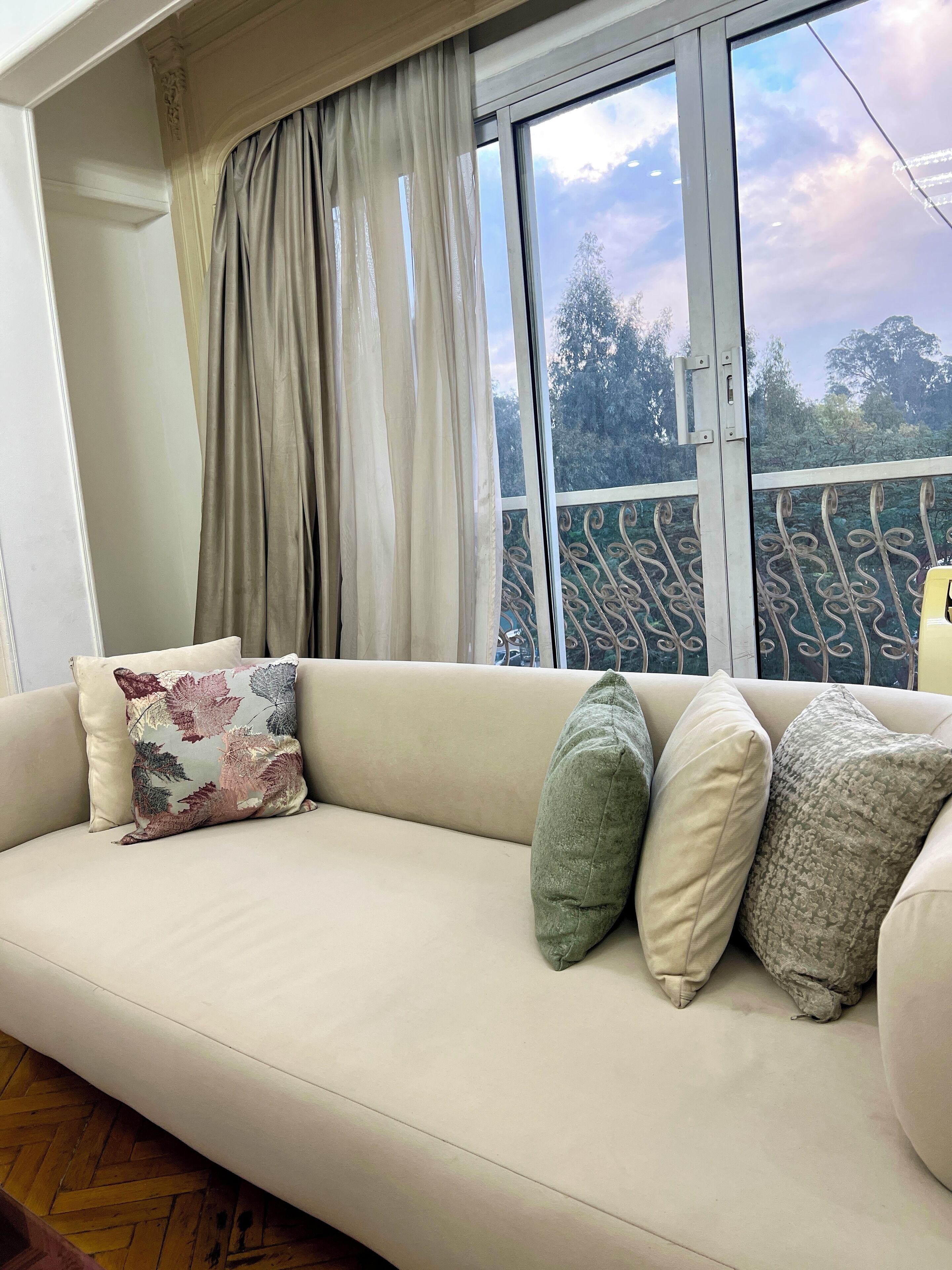 A beige sofa with patterned and solid pillows, a window with a view of trees, and a metal railing.