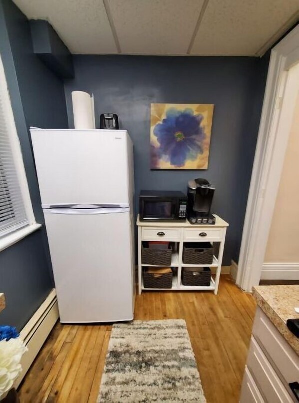 Compact kitchen with a refrigerator, microwave, and coffee maker, featuring a mounted artwork and wicker storage.