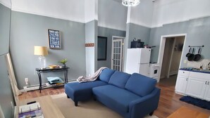 Een woonkamer met een blauwe bank, een houten salontafel en een eettafel. Er is een keuken met een koelkast en een magnetron.