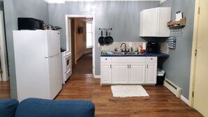 Een kleine keuken met witte kastjes, een fornuis en een magnetron. Op de voorgrond staat een blauwe bank.