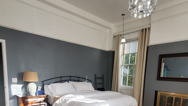 Een slaapkamer met een bed, nachtkastje, lamp, bankje en een raam met gordijnen.
