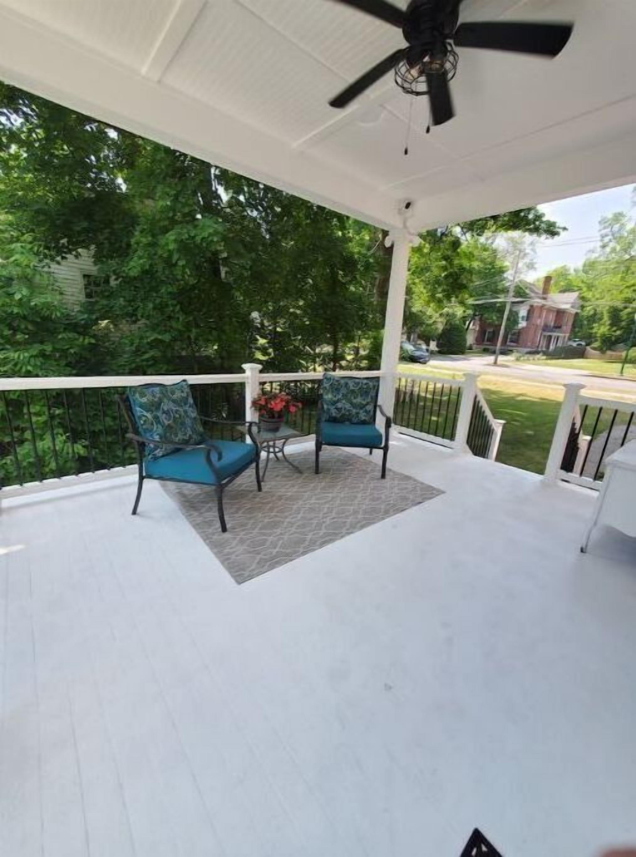 A covered porch with two blue chairs and a small table, surrounded by white railings and a white ceiling.