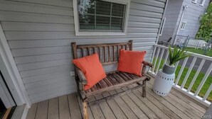 Un banc en bois avec des coussins orange, placé sur une terrasse en bois, une lanterne blanche et une fenêtre au-dessus.