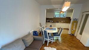 Un espai vital compacte amb un sofà gris, una taula de dinar amb cadires, una zona de cuina i una catifes de pell blanca sobre el terra de fusta.
