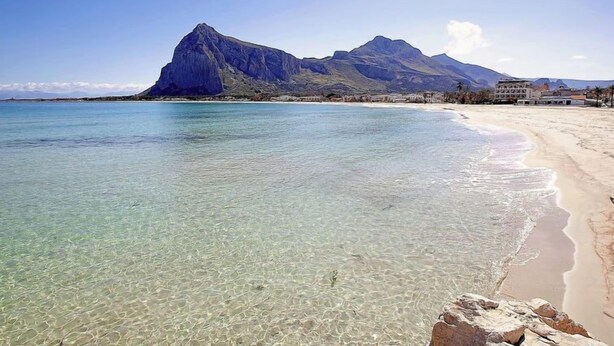 A beach with clear shallow waters, a prominent mountain in the background, and a few buildings along the shore. - Acquamarina Gaia by Interhome (San Vito lo Capo)