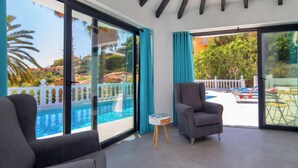 A modern room with a large sliding glass door leading to a pool area, a grey armchair, and a small table with a book. - Estrella Azul by Interhome (Calpe/Calp)