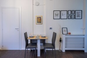 Un coin repas avec une table blanche, deux chaises noires et un banc blanc. Des tableaux encadrés ornent le mur, et un radiateur blanc se trouve en dessous.