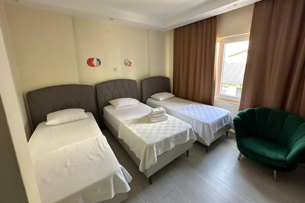 Habitación de hotel con tres camas individuales, un sillón verde y una ventana con cortinas.