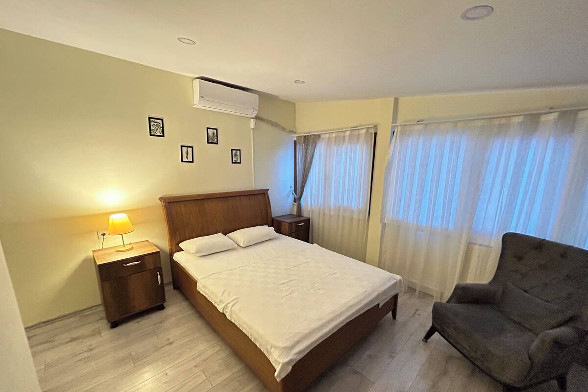 Una habitación de hotel con una cama de madera, una mesita de noche con lámpara, una silla y una ventana con cortinas translúcidas.