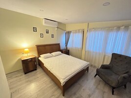 Una habitación de hotel con una cama de madera, una mesita de noche con lámpara, una silla y una ventana con cortinas translúcidas.